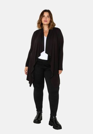 Zwarte draped cardigan van zachte stof, met een vloeiende vorm en open voorkant, gecombineerd met een wit shirt en zwarte broek, gedragen met chunky laarzen.