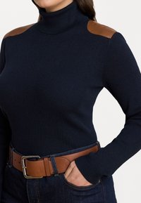Sini ribbed turtleneck neule, jossa on ruskeat nahkapartiontikäärmeet, yhdistettynä ruskeaan vyöhön tummansinisiin farkkuihin. Tasainen pinta, istuva muotoilu.