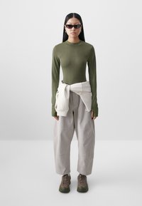 Haut ajusté vert olive à manches longues, associé à un pantalon beige ample et des baskets gris-vert. Un sweat-shirt blanc est noué à la taille.
