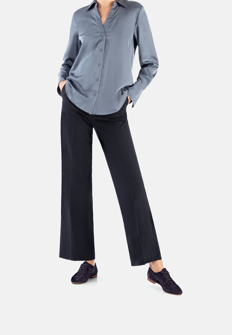 Femme portant un pantalon large foncé, une chemise bleue à manches longues boutonnée et des chaussures sombres à lacets, debout les mains dans les poches.