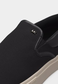 Μαύρο παπούτσι τύπου slip-on με υφασμάτινη επάνω επιφάνεια με υφή, μπεζ ελαστική σόλα και μικρό μεταλλικό λογότυπο "MK" στο πλάι κοντά στο άνοιγμα.