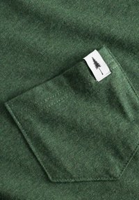 Donkergroene katoenen stof met een opgestikte zak en zichtbare stiksels, vergezeld van een klein wit label met een boomgraphic.