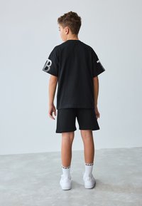 Sort bomuld t-shirt med korte ærmer, bagfra vist med et stiliseret bogstav på det ene ærme, kombineret med sorte shorts og hvide sokker.