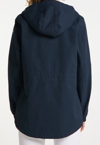 Mujer con una chaqueta con capucha de color azul oscuro, vista desde atrás, con mangas largas y un dobladillo ligeramente curvado sobre pantalones blancos.