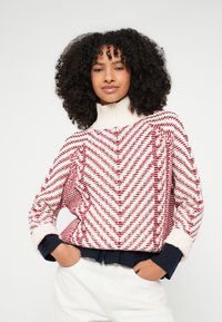 Πλεκτό πουλόβερ με λευκό και κόκκινο σχέδιο σε σχήμα ζιγκ-ζαγκ, ψηλό γιακά και χαλαρή εφαρμογή. Ριμπ λεπτομέρειες στις μανσέτες και στο κάτω μέρος σε ναυτικό μπλε.