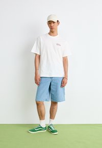 Hvit bomulls t-skjorte med teksten "Donut House", lyse denimshorts og grønne joggesko med hvite detaljer, båret med en beige caps.