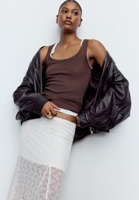 Brun ribbet tanktop lagdelt over en hvid bralette, kombineret med et gennemsigtigt hvidt blonderok og en oversized sort læderjakke. Guld ringdetalje.