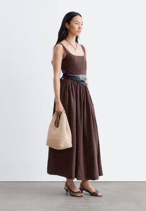Mujer con vestido maxi marrón sin mangas y pañuelo negro con estampado en la cintura, sosteniendo un bolso beige tejido, calzando tacones oscuros con puntera abierta.