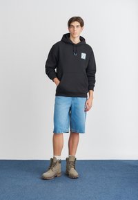 Sweatshirt preto com capuz e um bolso frontal, cordões azuis; combinado com calções de ganga azul claro e botas sociais bege.
