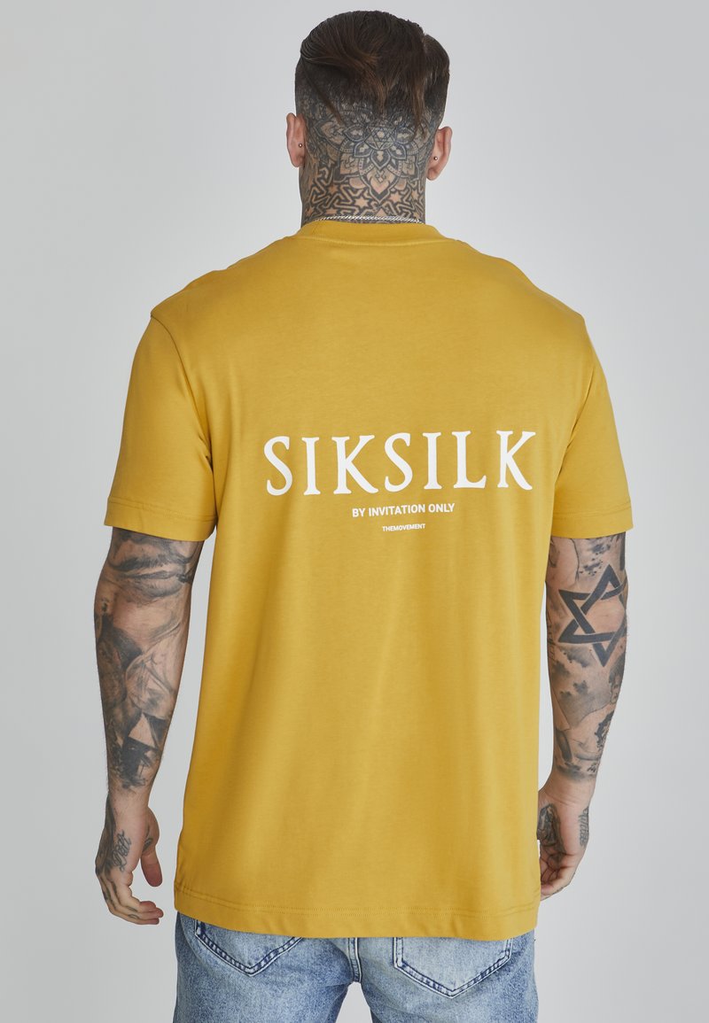 Žuta pamučna majica s kratkim rukavima i opuštenim krojem, sa "SIKSILK" logotipom otisnutim u bijelom na leđima. Nosite uz svijetloplave traperice.