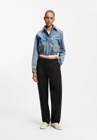 Κομμένο μπουφάν από μπλε τζιν με ελαστική ζώνη στη μέση και μανσέτες, συνδυασμένο με μαύρο παντελόνι ψηλής μέσης και εκρού αθλητικά παπούτσια.