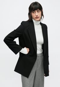 Tânără cu păr închis la culoare purtând un sacou negru, un guler înalt alb și pantaloni gri cu dungi subțiri, pe un fundal simplu, deschis.