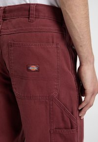 Pantaloni da lavoro color bordeaux realizzati in tessuto resistente, con cuciture rinforzate, più tasche e una patche quadrata sul retro.
