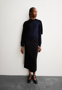 Marine blauwe button-up cardigan over een aansluitende zwarte rok. De outfit wordt compleet gemaakt met zwarte schoenen met decoratieve metalen accenten.