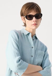 Μαύρα οβάλ γυαλιά ηλίου με σκούρους φακούς. Το μοντέλο φοράει ένα ανοιχτό μπλε πουκάμισο από λινό, με κουμπωτό μπροστά και γυρισμένα μανίκια.