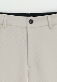 Un pantalon beige présentant une texture lisse, une fermeture à bouton et des passants de ceinture. La taille est sombre, contrastant avec le tissu clair.