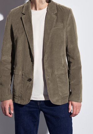 Man draagt een lichtbruine corduroy blazer over een wit t-shirt en donkerblauwe spijkerbroek, staand tegen een effen achtergrond.