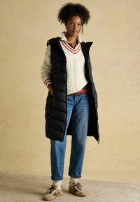 Czarna, długa kamizelka puchowa z kapturem, noszona na beżowym swetrze w warkocze z czerwonymi akcentami, w połączeniu z niebieskimi dżinsami i beżowymi trampkami.
