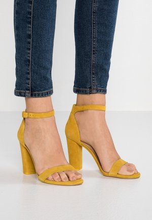 Füße, die gelbe Wildleder-Sandalen mit hohen Absätzen und Knöchelriemen tragen, kombiniert mit dunkelblauen Jeans, die oberhalb der Knöchel gekürzt sind.