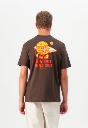 Homme portant un t-shirt marron avec un burger en cartoon orange tenant une planche de surf et le texte "SUN OUT BUNS OUT" au dos.