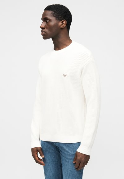 Witte gebreide trui met lange mouwen, geribde halslijn en manchetten, met een klein logo op de borst. Draagt blauwe spijkerbroek.