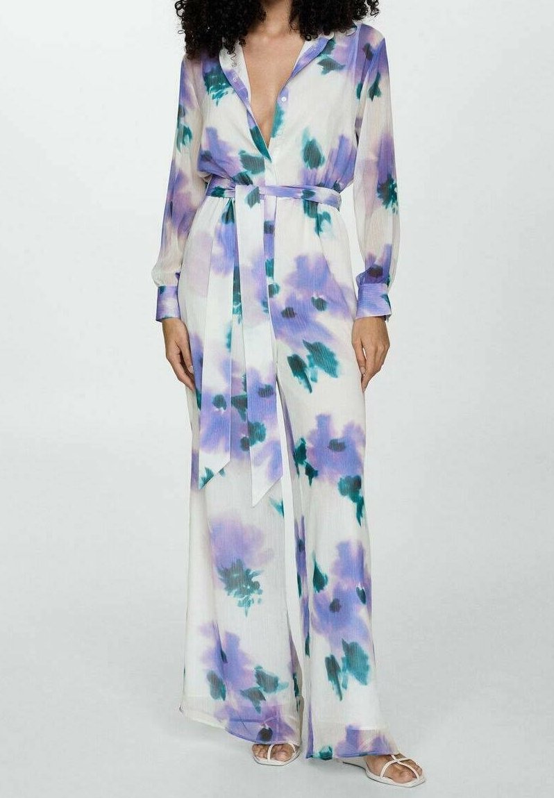 Combinaison blanche à manches longues avec imprimé floral violet et vert, taille ceinturée, décolleté profond en V et pantalon à jambes larges, portée avec des sandales blanches.