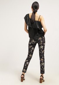 Mujer con el cabello oscuro recogido, vestida con un top negro de un solo hombro con volantes, pantalones ajustados de flores y sandalias negras de tacón alto, de pie de espaldas.