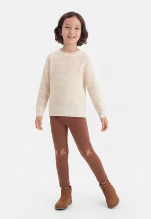 Kind glimlachend, draag een crèmekleurige gebreide trui, bruine legging met rendiergezichtjes op de knieën en bruine enkellaarsjes, staand op een witte achtergrond.