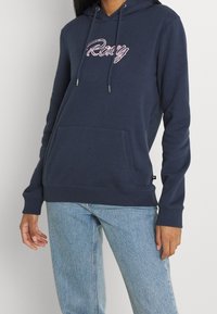 Sweat à capuche bleu marine en tissu doux, avec une poche kangourou avant et un logo "Roxy" brodé en rose sur la poitrine.