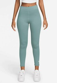 Kvinna i högmidjade teal leggings och vita sneakers, stående rakt mot en enkel vit bakgrund.