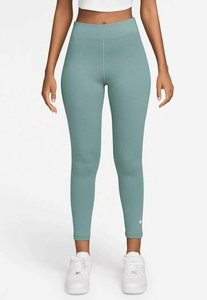 Kvinna i högmidjade teal leggings och vita sneakers, stående rakt mot en enkel vit bakgrund.