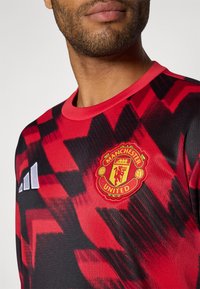 Červeno-černý dres s cikcakovým vzorem, kulatým výstřihem a logem Manchester United. Vyrobeno z lehkého a hladkého materiálu.