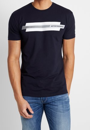 Mężczyzna ubrany w granatowy T-shirt z prostokątnym logo w paski białe i czarne z napisem "ANTONY MORATO", w połączeniu z niebieskimi dżinsami.