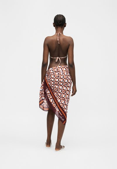 Γυναίκα με πλεξούδες, φορώντας μια φούστα με σχέδια και τοπ μπικίνι με λουράκι στον λαιμό, όρθια ξυπόλητη μπροστά σε ένα απλό φωτεινό φόντο.