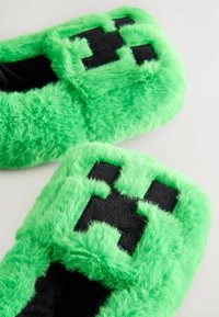 Chaussons verts duveteux avec une texture pelucheuse, ornés d'un design de visage pixelisé noir. Forme carrée avec un talon ouvert pour un enfilage facile.