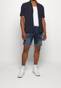 Lyhythihainen tummansinisestä kangasta valmistettu nappipaita, valkoinen toppi, farkkuhousut käärittyine helmoin ja valkoiset lenkkarit, henkilö seisomassa.