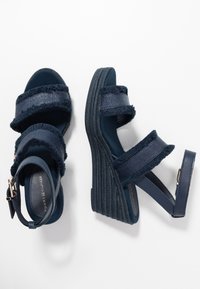 Sandali a zeppa blu navy con due cinturini in tessuto testurizzato e una cinghia alla caviglia. La suola presenta un design intrecciato in corda, che offre altezza e stabilità.