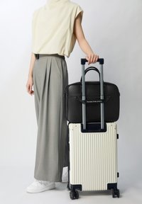 Bolso de cuero negro sobre una maleta blanca con textura estriada y asa retráctil. Modelo vistiendo una blusa crema, pantalones grises y zapatillas blancas.