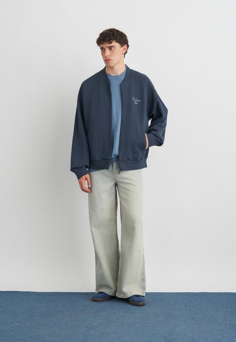 Young man wearing navy zip-up jacket, light blue shirt, wide light gray jeans, and navy sneakers, standing against a plain white wall.