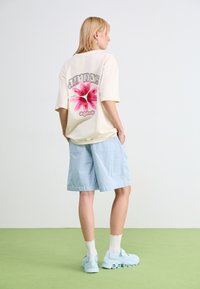 T-shirt in cotone crema con grafica floreale rosa e testo "Adidas Originals", abbinata a pantaloni corti a righe sottili blu chiaro e scarpe da ginnastica blu chiaro.