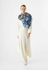 Écharpe en soie avec un motif floral en bleu, orange et marron, drapée sur une longue robe blanche avec des détails de boutons sur les manches.