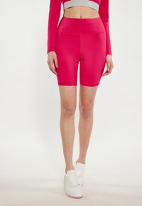 Pantaloni corti rosa aderenti con vita alta, realizzati in tessuto liscio e elasticizzato. Indossati con sneakers bianche con suola rosa.