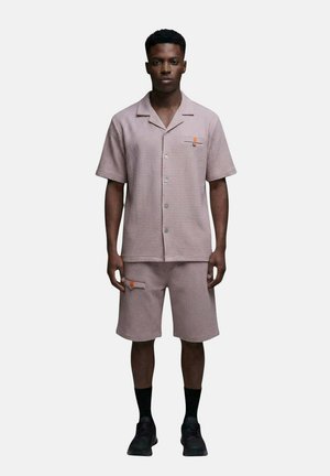 Homme portant une chemise à manches courtes boutonnée texturée violet clair et un short assorti avec des chaussettes noires et des chaussures noires, debout immobile.