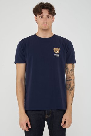Giovane uomo con capelli castani corti che indossa una t-shirt Moschino blu navy con un piccolo logo orsetto sul petto, mostrando il braccio destro tatuato.