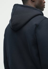 Tmavomodrá mikina s kapucňou, ktorá je veľká a mäkká, s hladkou textúrou a jednoduchým dizajnom. Pohľad sa zameriava na zadnú časť a oblasť ramien.