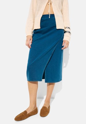 Niebieska spódnica z denimu w stylu owijanym, sięgająca do kolan z rozcięciem z przodu, w zestawieniu z beżowymi zamszowymi klapkami i krótkim swetrem w kolorze beżowym. Gładka tekstura, casualowy design.