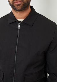 Giacca nera con colletto, zip frontale e due tasche anteriori. Realizzata in tessuto liscio senza motivi o accenti visibili.