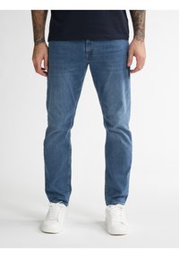 Jasnoniebieskie dżinsy o prostym kroju, z paskami na pasek i przednimi kieszeniami, zestawione z białymi trampkami na neutralnym tle.