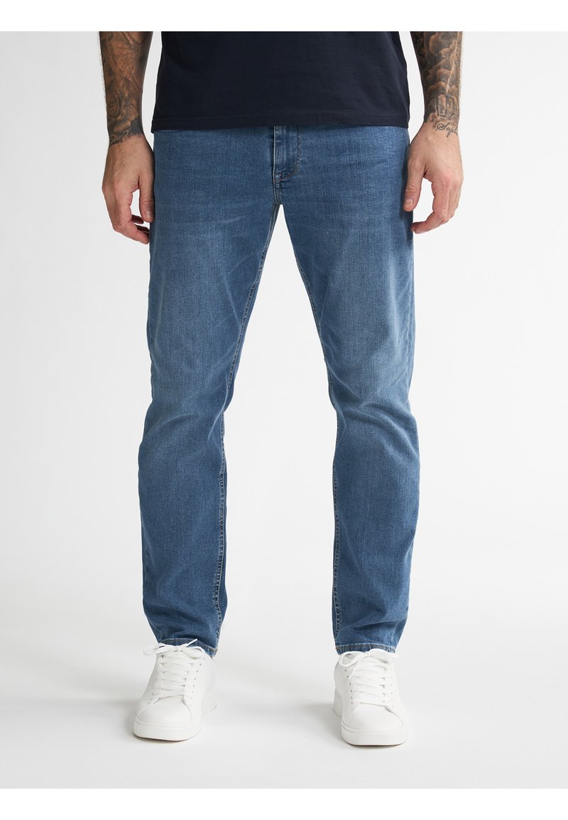 Jasnoniebieskie dżinsy o prostym kroju, z paskami na pasek i przednimi kieszeniami, zestawione z białymi trampkami na neutralnym tle.