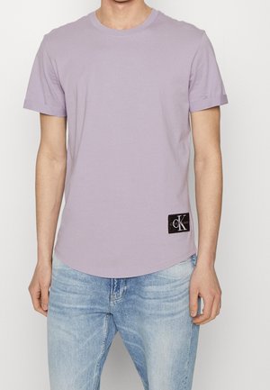 Person trägt ein hellviolettes T-Shirt mit kurzen Ärmeln und einem schwarzen Calvin Klein Logo-Patch nahe dem Saum sowie hellblaue Jeans.